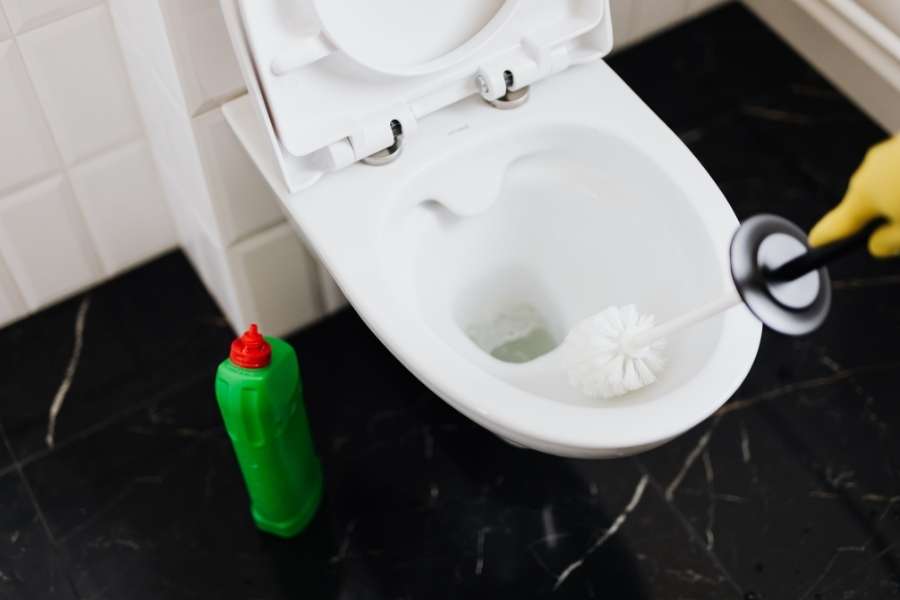 Une personne munie d'un gant de protection utilise une brosse de toilette pour nettoyer l'intérieur d'une cuvette blanche, avec une bouteille de produit nettoyant posée à côté.